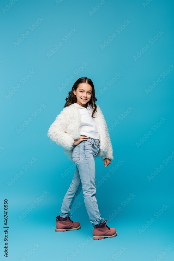full length of smiling preteen girl in faux fur jacket and winter boots standing with hand on hip on blue. full length of smiling preteen girl in faux fur jacket and winter boots standing with hand on hip on blue.
