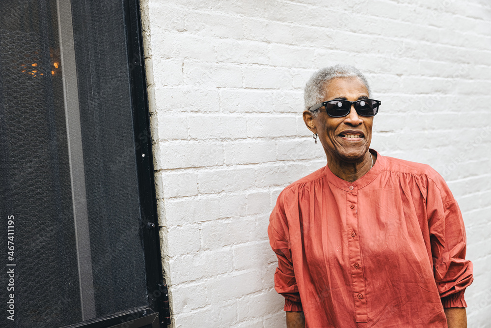 Obraz premium Senior black woman wearing sunglasses and smiling standing against a white brick wall