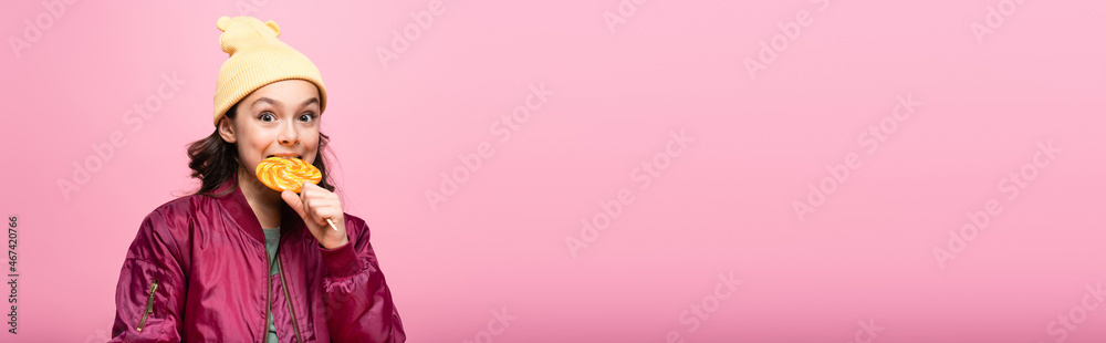 stylish preteen girl in winter outfit biting lollipop isolated on pink, banner. stylish preteen girl in winter outfit biting lollipop isolated on pink, banner.