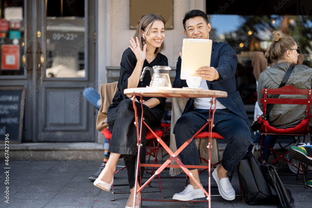 Smiling business people having video call on digital tablet at outdoor cafe. Concept of remote and freelance work. Idea of teamwork and business cooperation. Young european woman waving hand