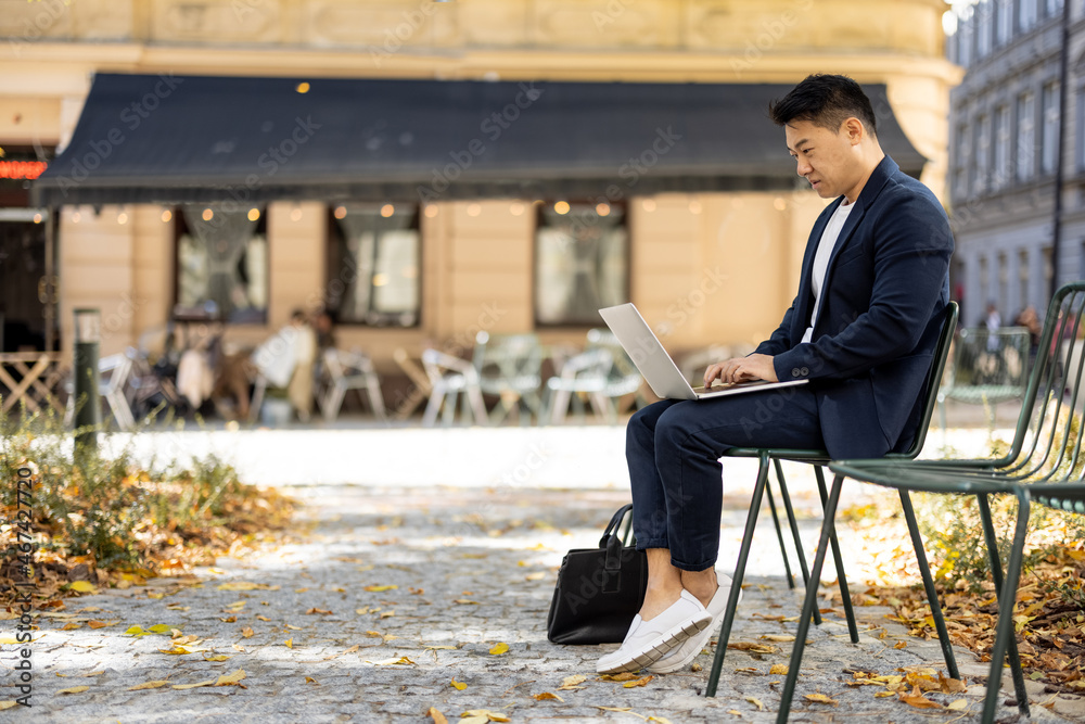 Fototapeta premium Focused asian businessman typing on laptop during work outdoors. Concept of remote work and freelance. Modern adult successful man wearing suit sitting on chair. Sunny autumn day