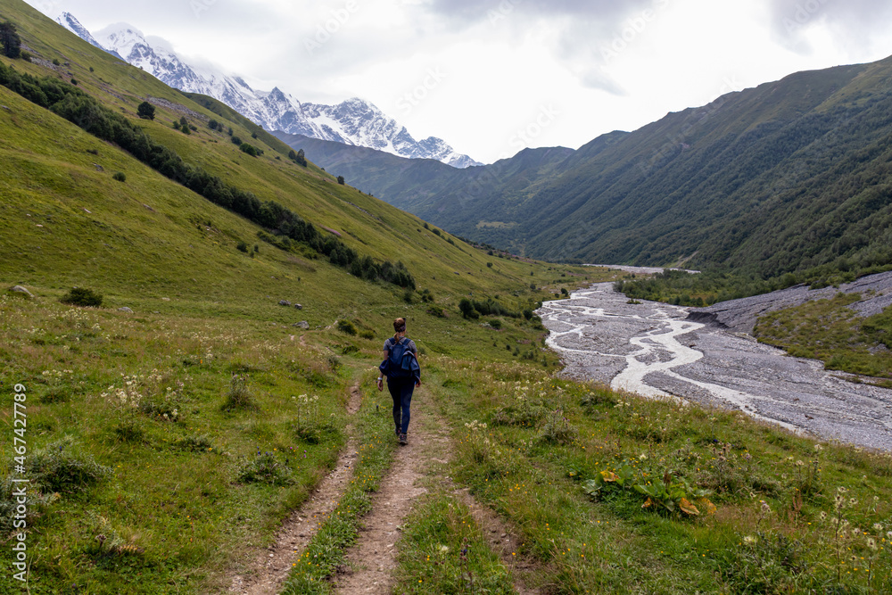 A woman hiking next to the river Adishischala with a view on the snow-capped peaks of Tetnuldi, Gistola and Lakutsia in the Greater Caucasus Mountain Range in Georgia, Svaneti Region. Wanderlust.