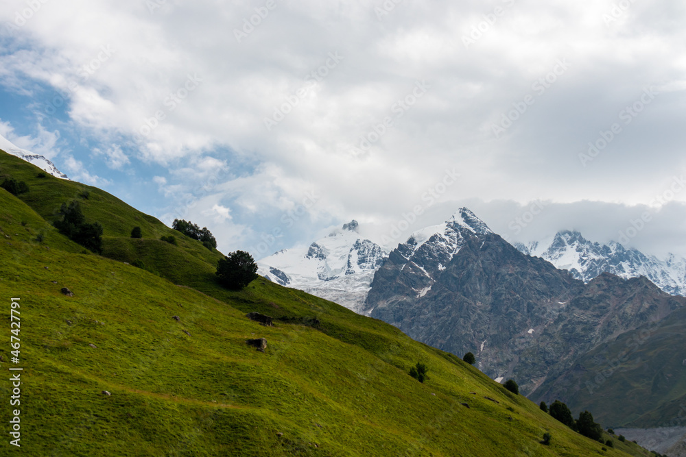 A panoramic view on the snow-capped peaks of Tetnuldi, Gistola and Lakutsia in the Greater Caucasus Mountain Range in Georgia, Svaneti Region. Hills with lush pastures, sharp peaks, hiking vibes.