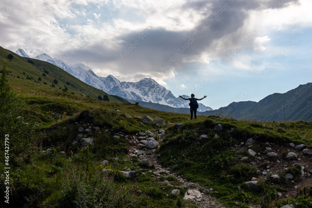 A woman enjoying the panoramic view on the snow-capped peaks of Tetnuldi, Gistola and Lakutsia in the Greater Caucasus Mountain Range in Georgia, Svaneti Region. Wanderlust, solitude, hiking trail.