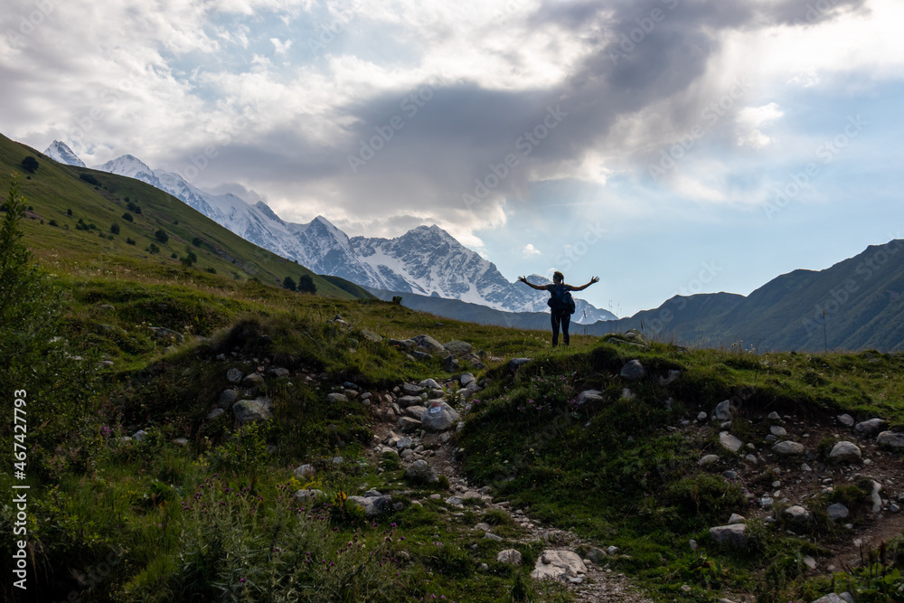 A woman enjoying the panoramic view on the snow-capped peaks of Tetnuldi, Gistola and Lakutsia in the Greater Caucasus Mountain Range in Georgia, Svaneti Region. Wanderlust, solitude, hiking trail. A woman enjoying the panoramic view on the snow-capped peaks of Tetnuldi, Gistola and Lakutsia in the Greater Caucasus Mountain Range in Georgia, Svaneti Region. Wanderlust, solitude, hiking trail.