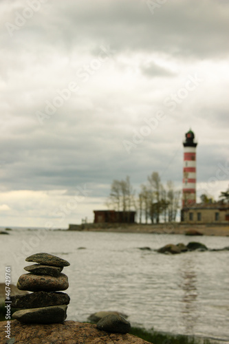 Pyramid made by stones. Stone tower and blurred lighthouse in the background. Sea, coastline and lighthouse. Beautiful seascape. Concept of balance, harmony and vacation. Rocks on the coast of the Sea