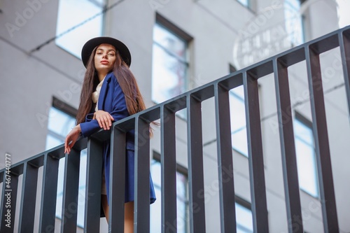 Fashion woman portrait - young pretty trendy girl posing at the city, autumn or spring street photo, classic black hat, soft smiling girl, trendy. Cold season. Blurred Details