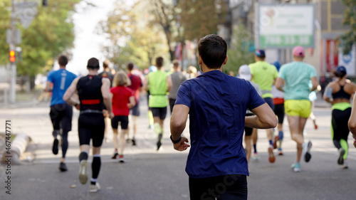 Man wearing blue t-shirt and earphones running on empty city road, blurred crowd of athletes running ahead. Contestants of sports event. Concept of healthy lifestyle