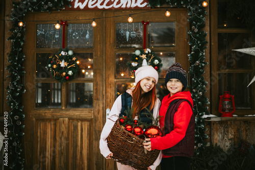 two children boy and teenage girl with stay on porch of village house with basket of toys and red balls, ready to decorate Christmas tree, during winter holidays, Christmas and New year vacation