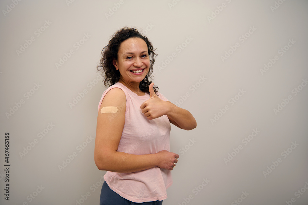 Portrait of African American happy woman smiling after getting a vaccine. Vaccinated woman showing thumb up and her arm with bandage after receiving immunity vaccine. Corona virus Vaccination concept Portrait of African American happy woman smiling after getting a vaccine. Vaccinated woman showing thumb up and her arm with bandage after receiving immunity vaccine. Corona virus Vaccination concept