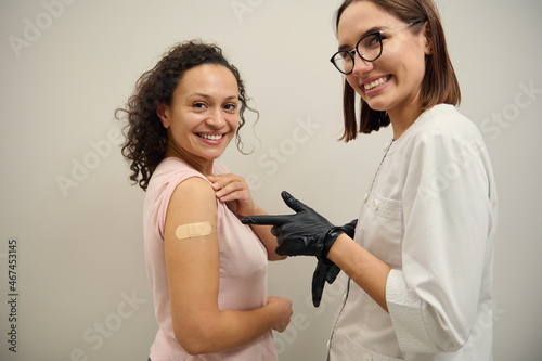 Cheerful nurse, medical professional, pointing to the hand of woman with band-aid after receiving corona virus or antiviral vaccine, both smiling looking at the camera. Voluntary vaccination concept