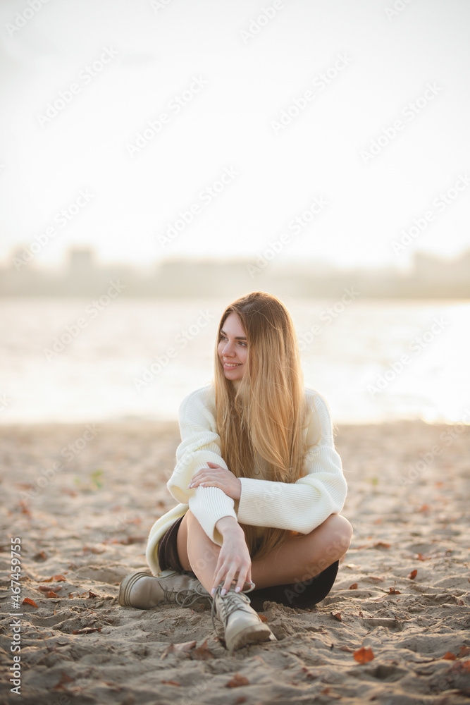 Portrait of a charming blonde with a gorgeous smile. A girl in a white sweater relaxes on the river bank