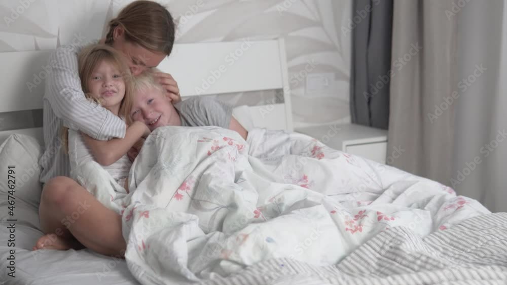 Mom hugs her kids in bed in the morning. A woman snuggles her son and daughter and kisses her beloved children. Mom hugs her kids in bed in the morning. A woman snuggles her son and daughter and kisses her beloved children.