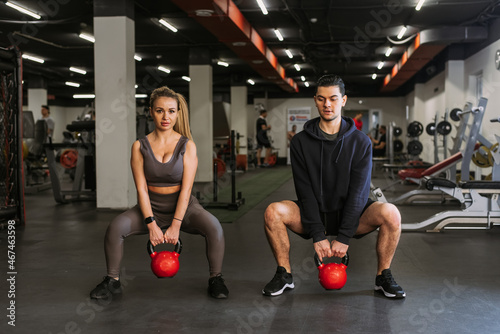 Fitness couple in sportswear squatting with weights and doing squats in the gym. A personal trainer in the gym corrects the squats of a young athlete