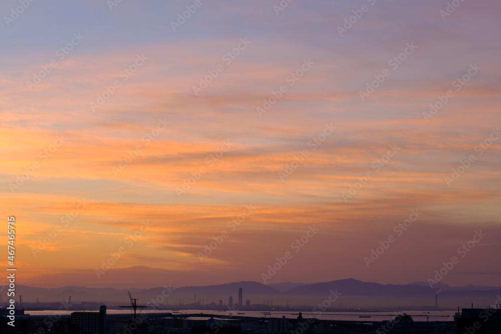 都市の夜明け。日の出とともに空と雲がオレンジ色に染まる。遠くに大阪湾を臨む