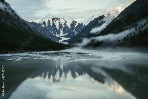 Scenic alpine landscape with snowy mountains in golden sunlight reflected on mirror mountain lake in fog among low clouds. Atmospheric highland scenery with low clouds on rocks and green mirror lake. © Daniil