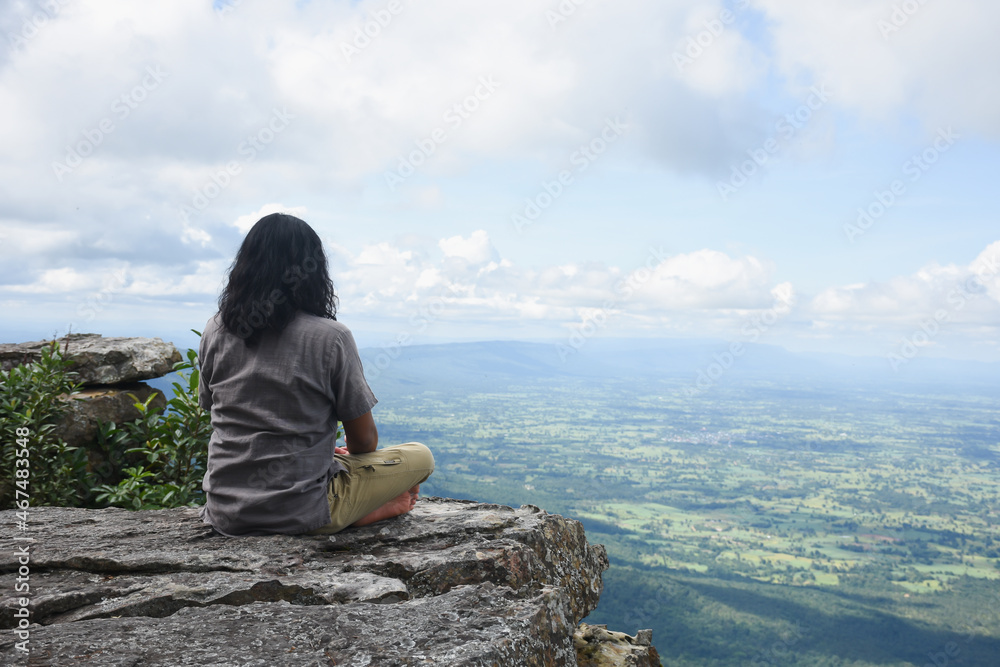 A man sits calmly on a cliff, looking at the view of the mountains and the clear sky.