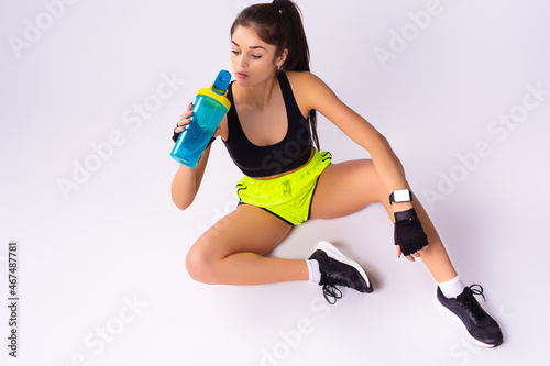 The pose of a pretty athletic girl sitting and drinking some water from the bottle in her hands after grueling workouts.
