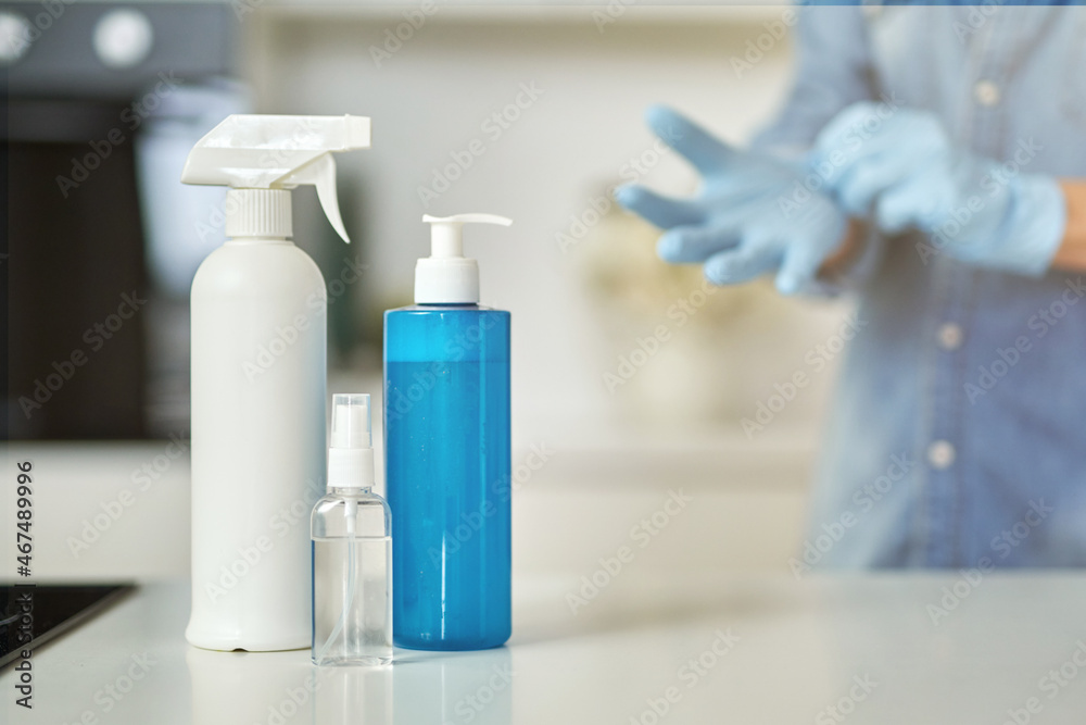 Sanitizer bottles to clean surfaces in the kitchen. Woman putting on protective gloves while getting ready for clean up in the background. Antiseptic, disinfection, cleanliness and healthcare Sanitizer bottles to clean surfaces in the kitchen. Woman putting on protective gloves while getting ready for clean up in the background. Antiseptic, disinfection, cleanliness and healthcare