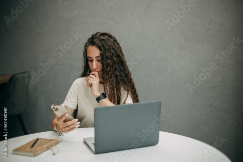 A young brunette girl with long wavy hair in a beige T-shirt works at home with a laptop, holds a smartphone in her hands, a notebook with a pen lies on the table