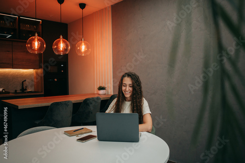 Smiling young brunette girl with long wavy hair in a beige T-shirt works at home behind a laptop, a notebook with a pen and a smartphone is on the table