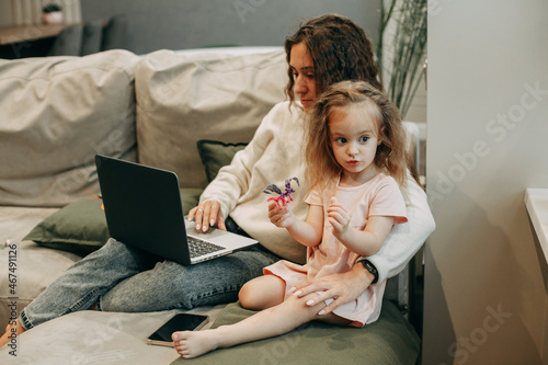 A young brunette woman with long wavy hair in jeans and a white sweater is working at home on the couch behind a laptop, a 2-year-old daughter with blond hair in a pink dress is playing next to