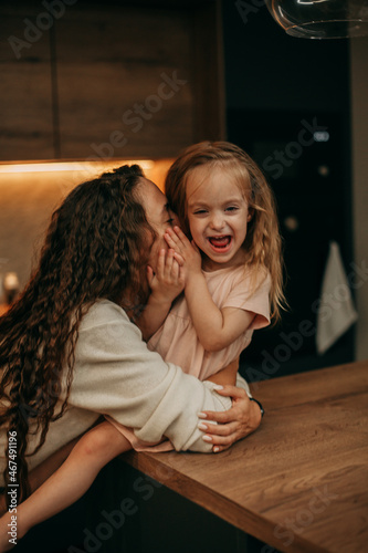 Happy brunette mom with long wavy hair in a white sweater hugs a smiling daughter 2 years old in a pink dress who sits on the kitchen bar table