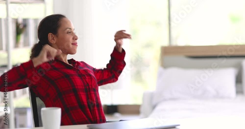 Glad ethnic woman in red checkered shirt closing laptop and stretching raised arms while working on remote project in sunlit bedroom at home