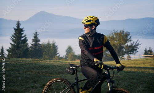 Fototapeta Naklejka Na Ścianę i Meble -  Close up of man in cycling suit riding bicycle on grassy hill. Male bicyclist in safety helmet enjoying the view of majestic mountains during bicycle ride. Concept of sport, bicycling and nature.