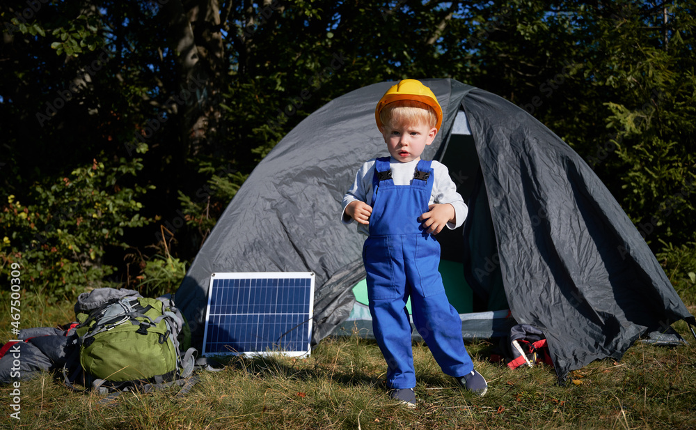 Portrait of focused little boy in construction helmet standing on grass in front of tent, one solar panel and backpack for hike. Young generation demonstrating new solar ecoenergy. Portrait of focused little boy in construction helmet standing on grass in front of tent, one solar panel and backpack for hike. Young generation demonstrating new solar ecoenergy.