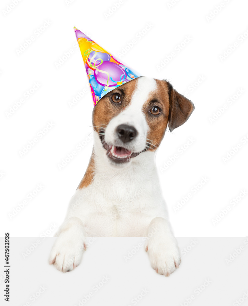 Happy jack russell terrier puppy wearing party cap looks above empty white banner. isolated on white background