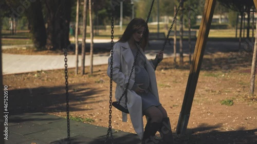 4K footage of a pregnant girl sitting on the swing in the park looks at the camera and then strokes her baby bump