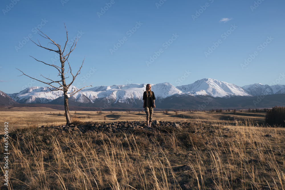 Woman and dry lonely tree in mountain valley. Snowy peaks of mountains on background, autumn yellow grass landscape Woman and dry lonely tree in mountain valley. Snowy peaks of mountains on background, autumn yellow grass landscape