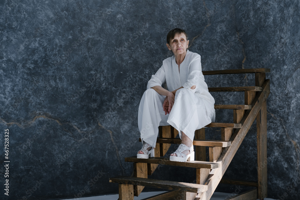 Studio portrait of beautiful looking old woman wearing white suit sitting on wooden stairs