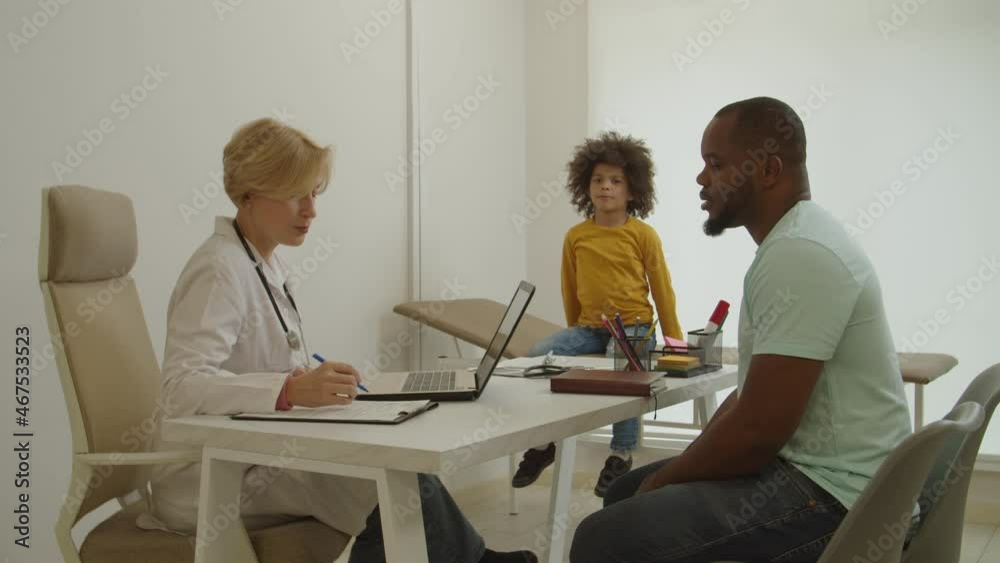 Positive attractive middle aged woman doctor communicating with caring black father and cute sick multiethnic elementary age boy about symptoms of illness, giving medical consultation at hospital. Positive attractive middle aged woman doctor communicating with caring black father and cute sick multiethnic elementary age boy about symptoms of illness, giving medical consultation at hospital.
