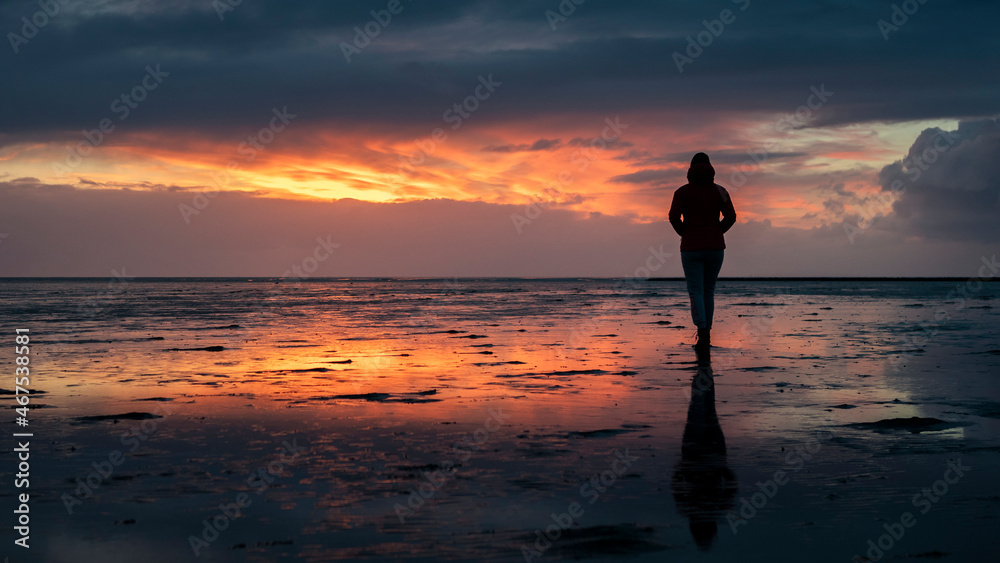 Woman silhouette walking along the Wadden Sea in Buesum during sunset, reflecting in the wet sand, Germany.
