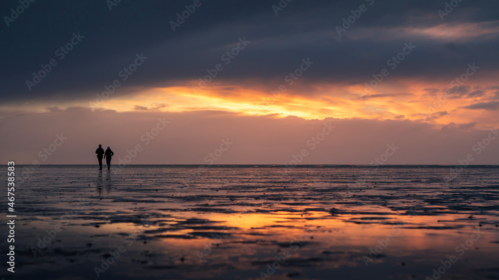 Couple silhouette walking along the Wadden Sea in Buesum during sunset, reflecting in the wet sand, Germany.