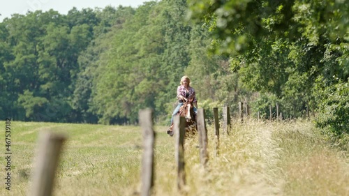 A cowgirl with blonde curls and a checked shirt rides a brown horse slowly along the edge of the forest in a field behind a fence.