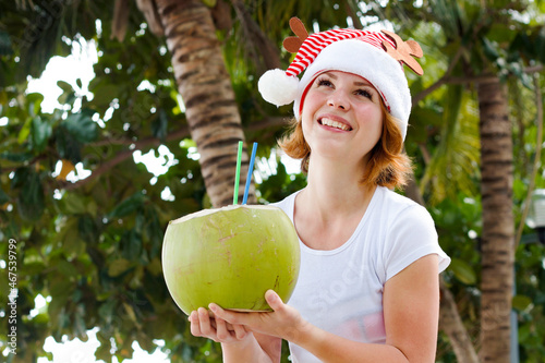 Young woman is enjoying the summer with a coconut drink in her hands and a coconut. Happy girl on the background of Malm in the red Santa hat celebrates the holiday. Merry Christmas Happy New Year photo