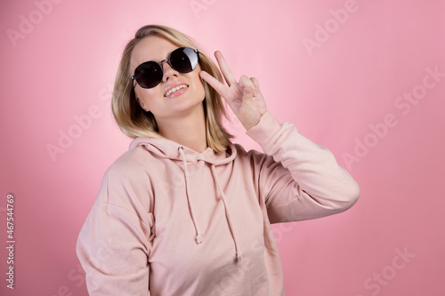 beautiful young woman with blonde hair wearing dark sunglasses makes a peace gesture, two fingers up, with one hand. In a pink hoodie on a pink background, studio portrait © Alexander