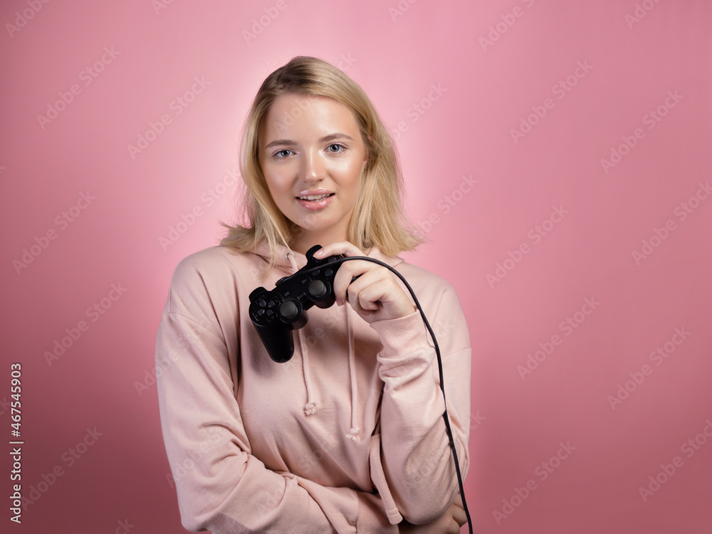 Charming blonde gamer. A young smiling woman in a pink hoodie holds a joystick from a game console in her hands, winning the round. Portrait on a pink background