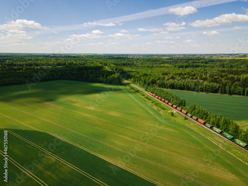 Freight train heading to the green forest in summer from above. Fields and crops. 