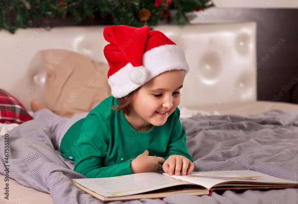 Little cute girl in Santa cap and Christmas pajamas reading a book. Little cute girl in Santa cap and Christmas pajamas reading a book.