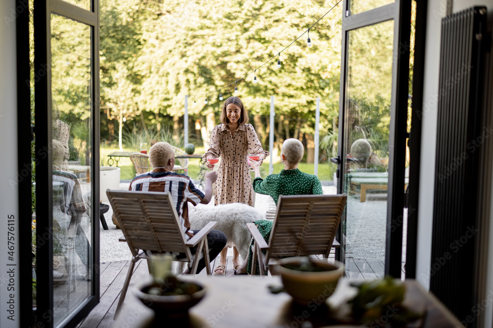 Woman giving cocktails to a friends sitting on wooden chairs at home terrace. European girls and latin man spending time together. Concept of leisure. Modern domestic lifestyle Woman giving cocktails to a friends sitting on wooden chairs at home terrace. European girls and latin man spending time together. Concept of leisure. Modern domestic lifestyle