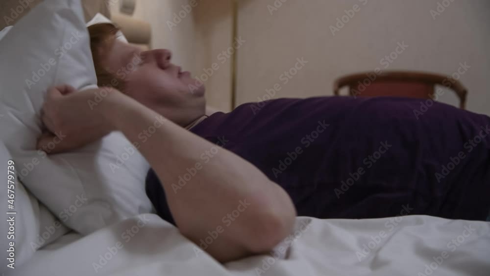 Close-up side view of exhausted adult redhead caucasian man in purple t-shirt lying on bed with white linens and trying to sleep. Man straightens the pillow several times. Uncomfortable bedding theme. Close-up side view of exhausted adult redhead caucasian man in purple t-shirt lying on bed with white linens and trying to sleep. Man straightens the pillow several times. Uncomfortable bedding theme.