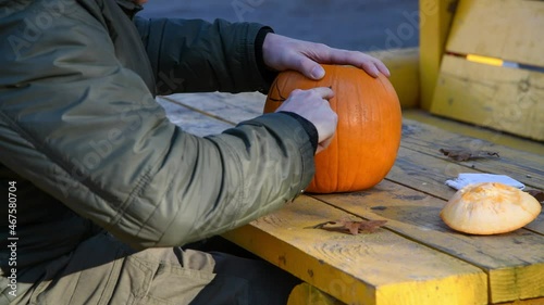 Side view of caucasian man sitting at yellow table in gazebo and carving Jack O'Lantern face on orange Halloween pumpkin. Person wears green clothes. Holiday handmade decorations theme. photo