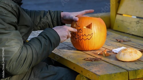 Side view of caucasian man sitting at yellow table in gazebo and carving Jack O'Lantern face on orange Halloween pumpkin. Person wears green clothes. Holiday handmade decorations theme. 4K resolution. photo