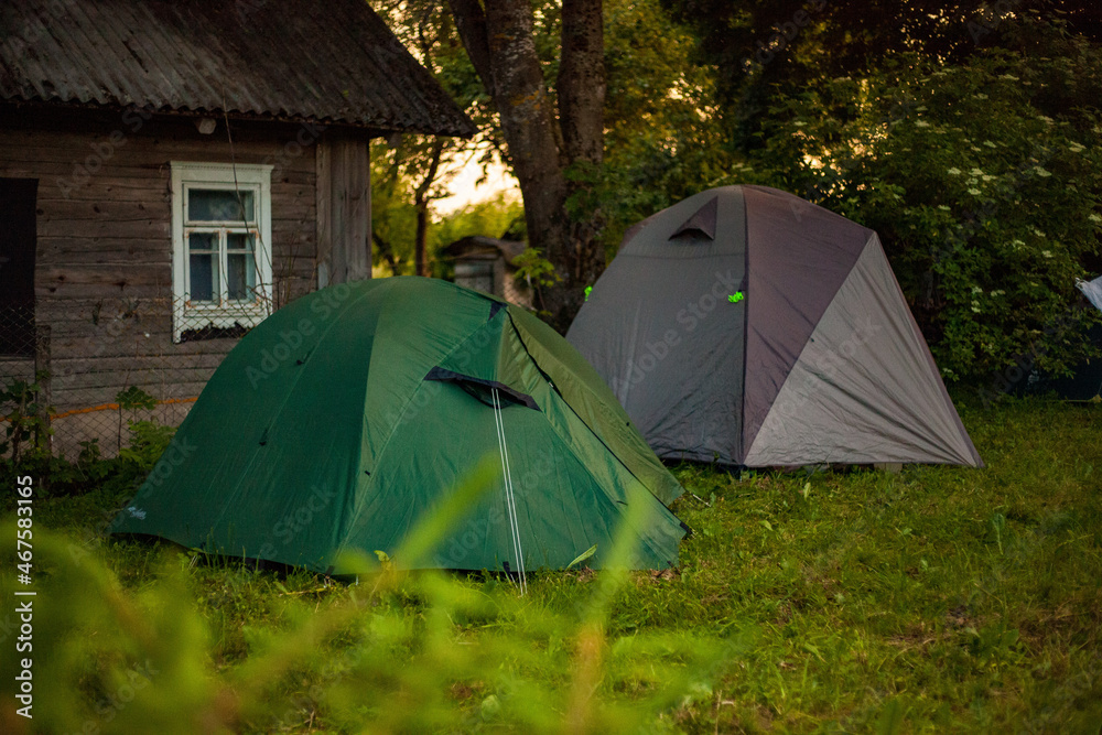 Photo of two tents near a wooden house on green grass in a village outside the city in nature