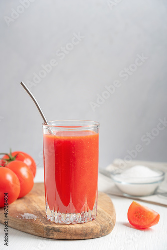 Tomato juice made from ripe red tomatoes, usually used as a beverage, either plain or in cocktails, served in tall drinking glass with reusable metal straw on white wooden table with salt. Vertical