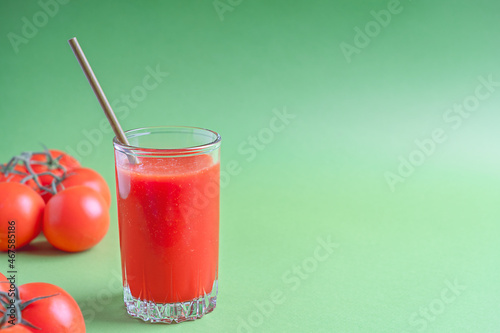 Portion of organic tomato juice made of blended ripe red tomatoes served in tall drinking glass with paper straw against bright green background with ingredients. Image with copy space, horizontal